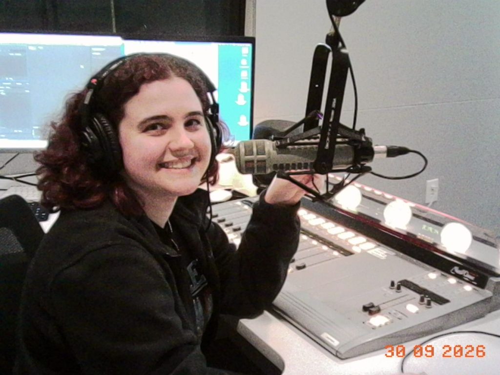 "Isabel in Studio"
Co-host Isabel smiling to the camera sitting at a desk in front of the audio mixer, with headphones on, holding onto the microphone.